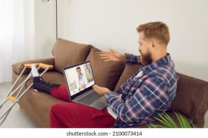 Man With Leg Injury While Sitting At Home Talking To Doctor On Video Call During Online Consultation. Adult Bearded Man With Bandage On His Leg Sitting On Couch With Laptop, Crutches Nearby.