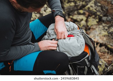 A man leisurely sits on a rock, packing his backpack for an adventure. Soil beneath his feet, he gestures with his thumb as he prepares his luggage and bags, including a helmet - Powered by Shutterstock