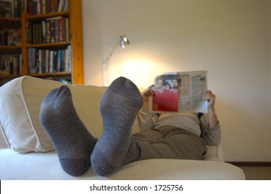 A Man Lies On The Couch With His Feet Up, Reading The Paper. Focus On The Slightly Worn Socks. Space For Text On The Wall, Top Right.