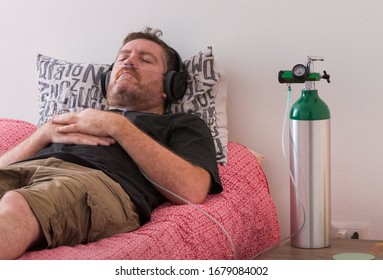A man lies on his bed while breaths through a cannula connected to an oxygen cylinder