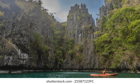 A man in a life jacket is sailing a canoe on the emerald lagoon. Sheer karst cliffs with green vegetation on steep slopes surround the bay. Blue sky, clouds. Philippines. Palawan.Small lagoon.  - Powered by Shutterstock