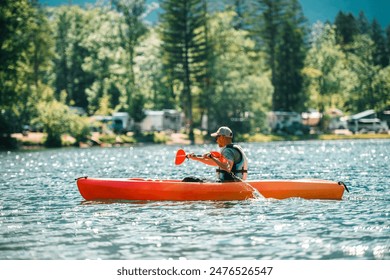 A man in a life vest is paddling a red kayak on a calm lake. The water is sparkling, and the lake is surrounded by a forest of tall trees. There are a few motorhomes and vehicles parked on the shore. - Powered by Shutterstock