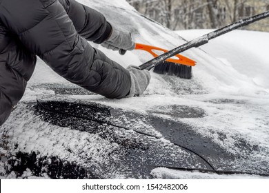 Man Lifted The Windshield Wiper To Clear The Snow From The Windshield, Using Car Snow Brush For This. Person In A Black Jacket And Gray Gloves.  