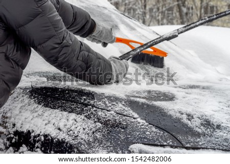 Man lifted the windshield wiper to clear the snow from the windshield, using Car Snow Brush for this. Person in a black jacket and gray gloves.  