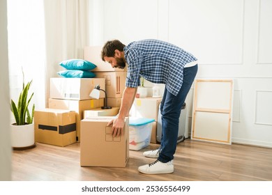 Man lifting a heavy box while moving into a new apartment, facing challenges of unpacking furniture and settling in, determined to create a cozy space despite back pain and feeling alone