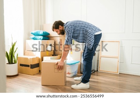 Man lifting a heavy box while moving into a new apartment, facing challenges of unpacking furniture and settling in, determined to create a cozy space despite back pain and feeling alone