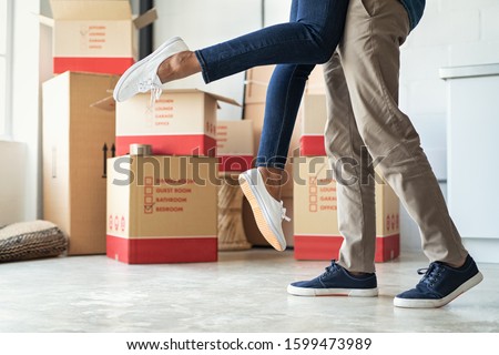 Man lifting his girlfriend among cardboard boxes as they start living together in new house. Closeup of excited husband embracing wife celebrating relocation. Loving man and woman standing near boxes.