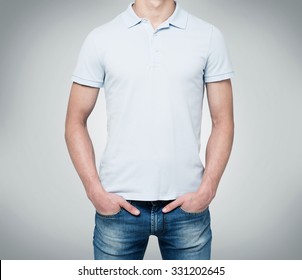 A man in a light blue polo shirt and denims holds his hands in pockets. Grey background.