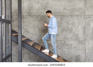 A man in a light blue shirt and jeans walks up a modern concrete staircase while looking at his smartphone. - Powered by Shutterstock