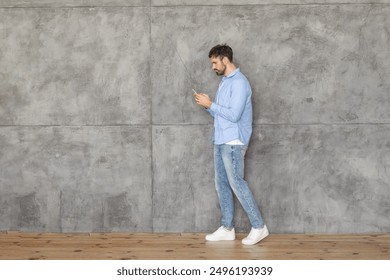 A man in a light blue shirt and blue jeans walks past a concrete wall while looking at his smartphone. He is wearing white sneakers. The floor is wooden, copy space - Powered by Shutterstock