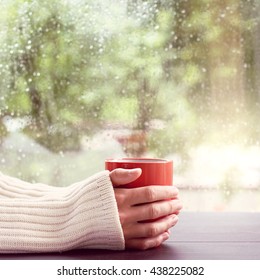 Man In A Light Jumper, Holds In Hands Red Mug On A Background Of A Wet Window After The Rain / Hot Drink And Watch The Rain