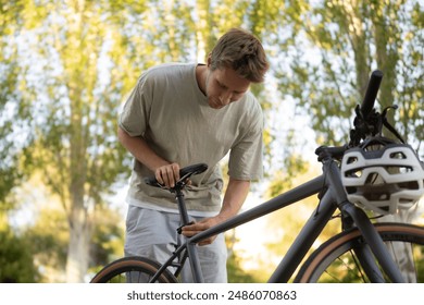 man in a light-colored shirt adjusts the seat height of his bicycle. He is standing outside on a sunny day - Powered by Shutterstock