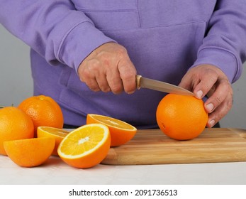 A Man In A Lilac Sweatshirt Cuts Oranges In Half To Squeeze Orange Juice. Close-up Of Hands, Oranges, Knife And Wooden Board