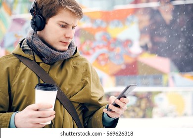 Man listening to music with headphones looking to the phone and drinking coffee in the winter on the street. Concept of technology and lifestyle - Powered by Shutterstock