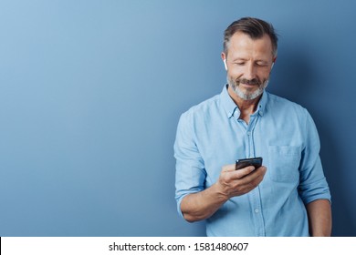 Man Listening To Music Or Media On His Mobile Phone Using An Ear Bud Posing Over A Blue Studio Background With Copy Space