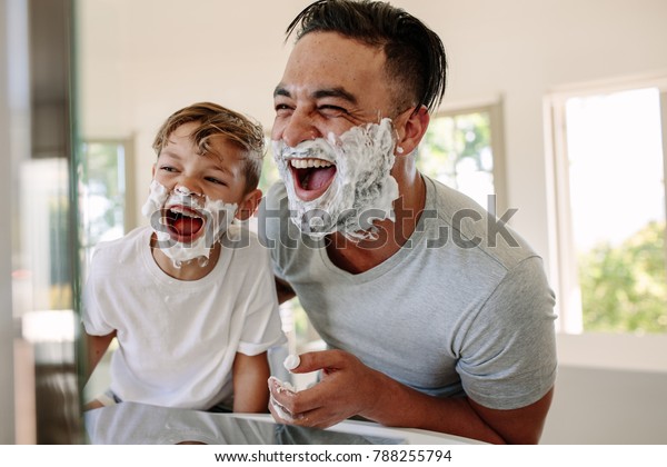 Man and little boy with shaving foam\
on their faces looking into the bathroom mirror and laughing.\
Father and son having fun while shaving in\
bathroom.