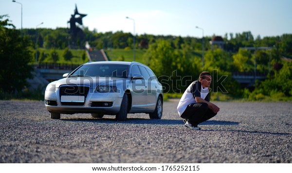 Man with Lonely silver car\
in an empty car gravel area in summer park. Guy squatting near\
automobile