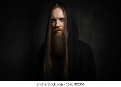 Man with long hair and beard in black hood on dark background