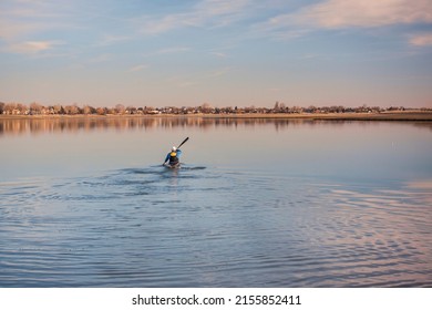man in a long racing kayak with a wing paddle is paddling away on a calm lake leaving waves and splashes, early spring scenery in Colorado