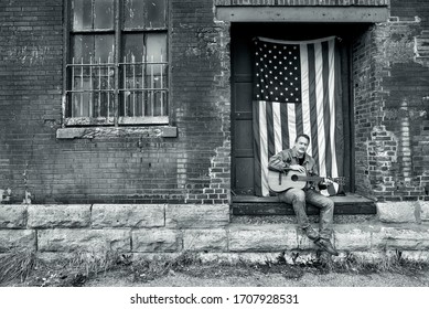 Man Looking At Camera Sitting On Brick Factory Loading Dock In Jeans With Acoustic Guitar And American Flag, Freedom, Country, Usa, Independence, Playing Guitar Outside, Singing, Portrait, Music