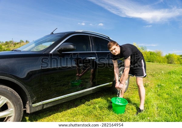 Man Looking at Camera and
Wringing Out Soapy Sponge into Green Bucket While Washing Black
Luxury Vehicle in Green Grassy Field on Bright Sunny Day with Blue
Sky