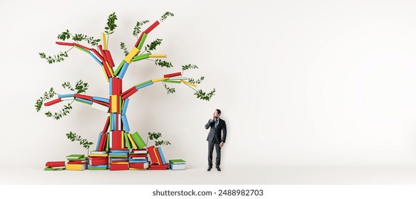 A man looking at a colorful tree sculpture made of books with green leaves, set against a white background, depicting the concept of growth through knowledge - Powered by Shutterstock