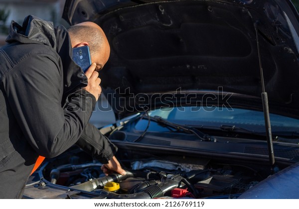 man looking at the engine of the broken car and\
calling for help with the\
phone