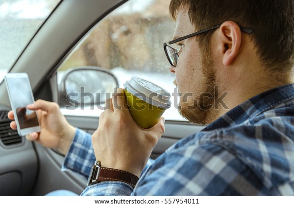 man looking info in phone and drinkig coffie while\
sitting in car