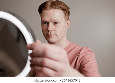 Man Looking into Vanity Mirror Preparing for Skincare Routine
A young man looks thoughtfully into a vanity mirror with LED lighting, preparing for a skincare or grooming routine. 
sstkDiverseBeauty