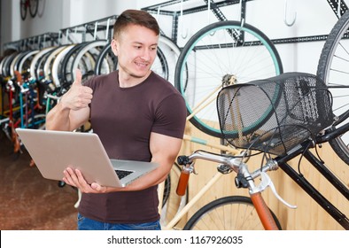 Man Is Looking In Laptop And Choosing Modern Bicycle In The Bike Store.