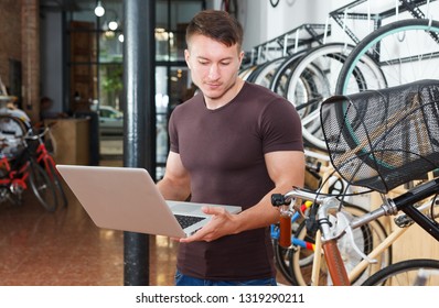 Man Is Looking In Laptop And Choosing Modern Bicycle In The Bike Store.