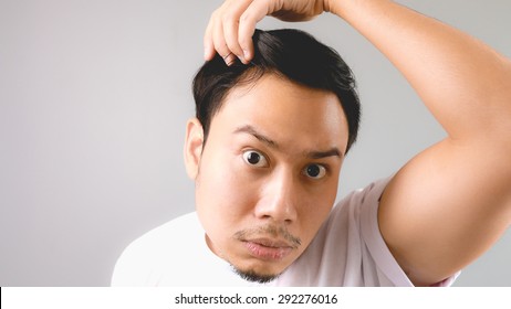 A man looking at the mirror and shocked that he is losing his hair. An asian man with white t-shirt and grey background.