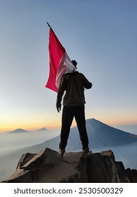 a man looking at the mountain above the clouds holding the Indonesian flag as the sun rises over a rock cliff