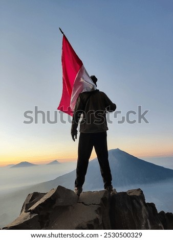 a man looking at the mountain above the clouds holding the Indonesian flag as the sun rises over a rock cliff