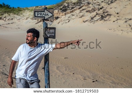Man looking to one side and point to the other by the side of signs with the sayings Beach / Trail in portuguese in sand dunes
