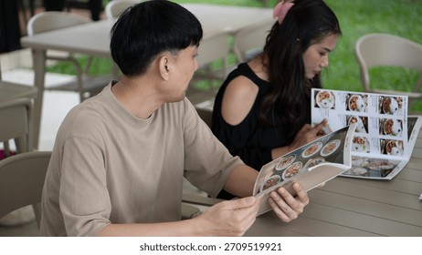 Man looking at a restaurant menu while sitting with a woman at an outdoor cafe, selecting a meal.