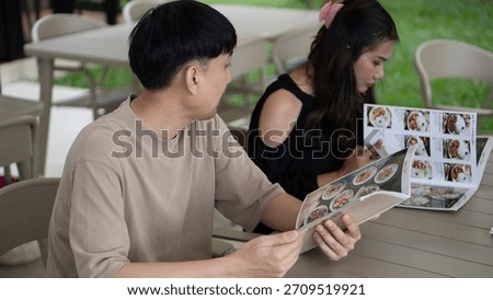 Man looking at a restaurant menu while sitting with a woman at an outdoor cafe, selecting a meal.
