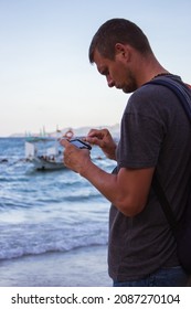 Man Looking At Smartphone On Seacoast. Handsome Man With Telephone On Seaside With Boat On Background. Tourist In Tropics. Serious Tourist With Phone. Leisure On Island. Winter Vacations In Asia.