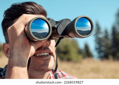 Man looking through binoculars outdoors on sunny day. Mountain landscape reflecting in lenses