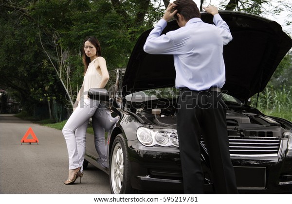 Man looking under car hood of broken down car,
woman waiting