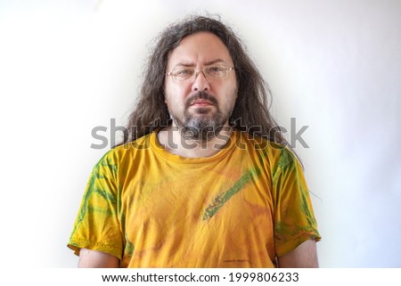 The man looks forward, squinting intently. A casually and sloppy dressed man with a beard and long hair in a paint-stained T-shirt on a light background.