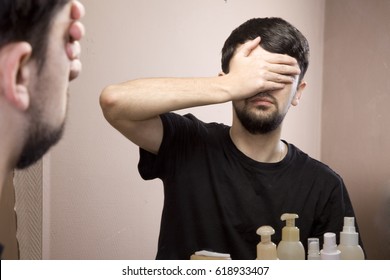 The man looks in the mirror. A man is worried about his appearance. Feeling of inferiority, complexes, low self-esteem. He covers his face with his hands - Shutterstock ID 618933407