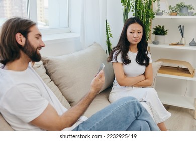 A man looks at the phone screen during an argument with his girlfriend. The angry and hurt woman looks in his direction and is sad. Family discord at home, phone addiction - Shutterstock ID 2232925419