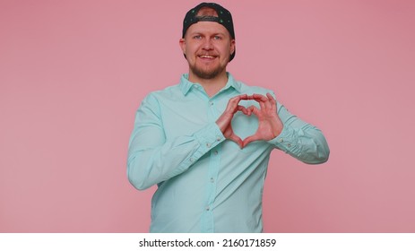 Man In Love. Smiling Man 30s In Blue Shirt Makes Heart Gesture Demonstrates Love Sign Expresses Good Feelings And Sympathy. Young Adult Blonde Guy Boy Isolated Alone On Pink Studio Wall Background