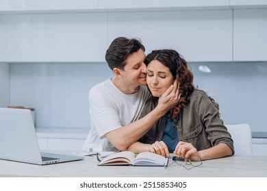 Man lovingly comforting stressed woman working from home while sitting at desk with laptop and notebook in cozy kitchen environment. Couple in love taking care of each other - Powered by Shutterstock