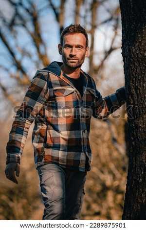 Man in lumberjack clothing is moving around a tree, looking into camera