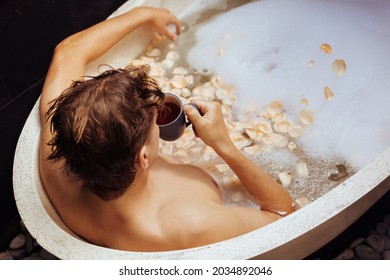 Man lying in bath tub with soap foam, petals, mug