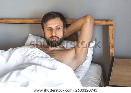 Man lying in bed with pensive expression, hand behind head, reflecting silently in minimalist bedroom, concept of midlife crisis and self-reflection