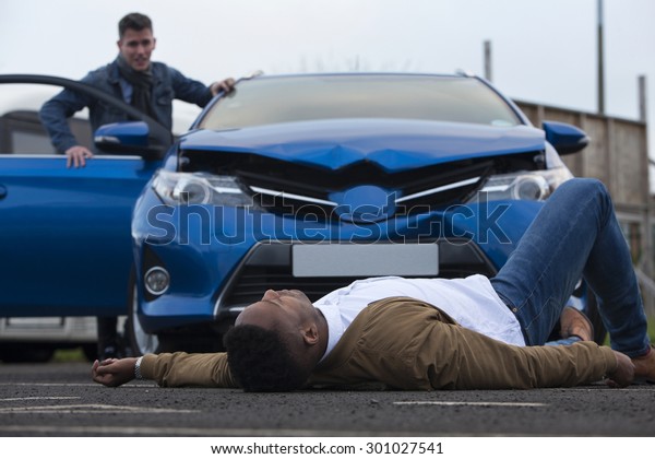 A man is lying in front of\
a damaged car which has just hit him. The responsible driver is\
standing at the door of the car, looking at him with a worried\
expression.