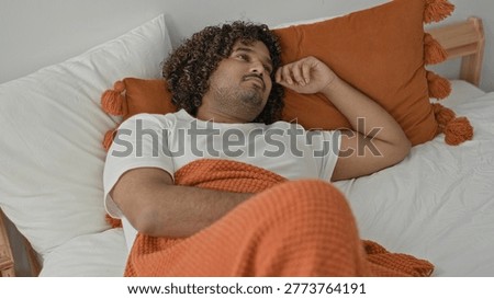 Man lying on bed with hand to temple, orange pillow and knitted blanket visible in building; sleepy repose.